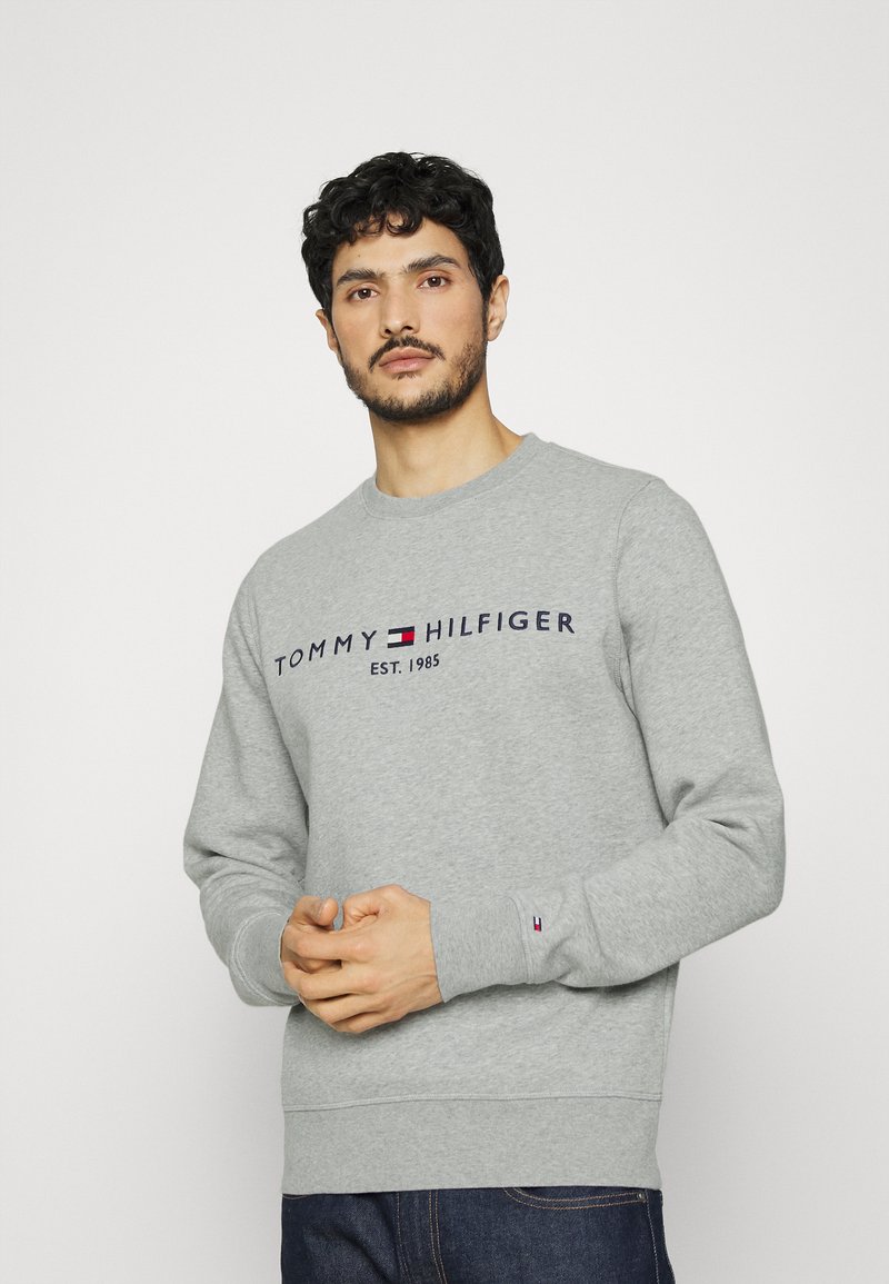 Sudadera gris hecha de tela suave, con cuello redondo, mangas largas y un logotipo con acentos rojos y azules en el pecho.