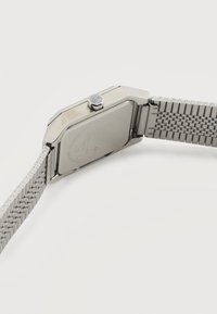 Ασημένιο μεταλλικό ρολόι χειρός με μπρασελέ από δίχτυ, εμφανιζόμενο από πίσω, που τονίζει την κορόνα και το τετράγωνο κουτί του ρολογιού σε απλό λευκό φόντο.