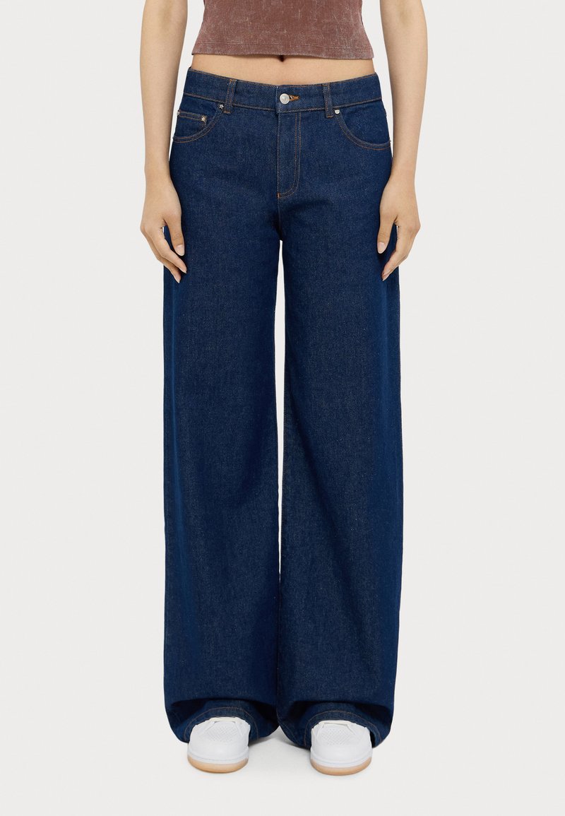 Jean en denim à jambes larges de couleur bleu foncé, avec un design classique à cinq poches, des coutures orange et une fermeture à bouton à la taille.