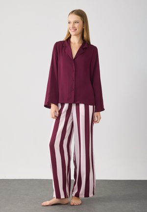 Donna sorridente in piedi a piedi nudi, con una maglietta a maniche lunghe bordeaux con bottoni e pantaloni a gamba larga a righe verticali rosa e bordeaux.