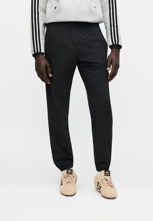 Osoba ubrana w czarne spodnie dresowe z elastyczną talią, beżowe buty sportowe Adidas oraz szarą bluzę z czarnymi paskami na rękawach.