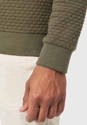 Olivgrüner strukturierter Pullover mit einem gewebten Muster, gerippten Bündchen und Abschlüssen, dargestellt neben einer Hand vor einem hellen Hintergrund.