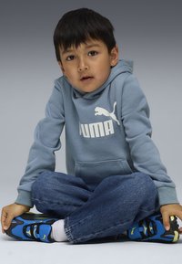 Sweat �à capuche bleu en polaire avec logo blanc, jean en denim et chaussures de sport bleues. Enfant assis en tailleur, fond neutre.