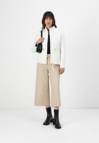 Gesteppten weißen Jacke mit beigen Ärmeln, schwarzem Oberteil, beigen weit geschnittenen Hosen und schwarzen Absatzstiefeln. Hält eine kleine schwarze Handtasche.