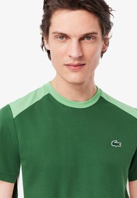 Tricou verde cu mânecă scurtă, dintr-un material texturat, având o secțiune de umăr de un verde mai deschis și un mic logo brodat pe piept.