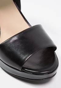 Sandale à talons hauts ouverte en cuir lisse de couleur noire, avec une large bride à l'avant et une finition brillante sur la semelle.