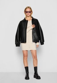 Svart läder oversized bomberjacka, beige knäppt klänning och svarta chunky anklarstövlar. Modell som bär kattöga solglasögon, stående mot en grå bakgrund.