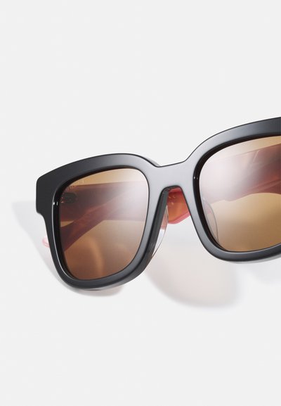 Lunettes de soleil noires surdimensionnées avec des verres dégradés marron, présentant une monture géométrique et des accents rouges contrastants sur les côtés.