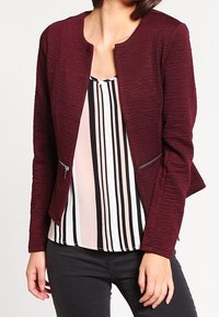 Veste texturée bordeaux avec un col rond, des poches latérales zippées et une coupe ajustée, portée sur un haut rayé noir et blanc.