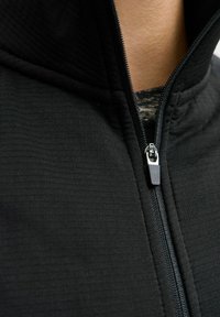 Sudadera negra con cremallera, que presenta un tejido texturizado, un cuello alto y una cremallera lisa con una pequeña lengüeta para facilitar el cierre.
