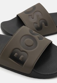 Mørkebrune sandaler med sklisikker såle i teksturert svart og en bred stropp med preget logo i matchende brunt.