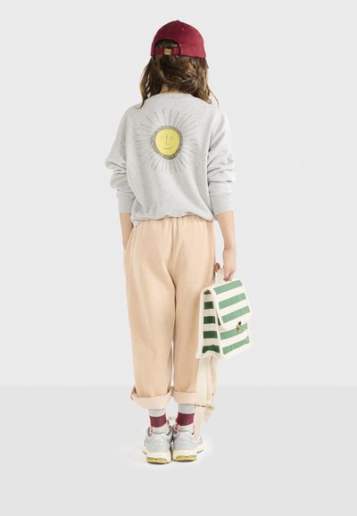 Sudadera gris con un gráfico de sol, pantalones beige de canalé, zapatillas blancas con detalles amarillos, llevando una bolsa a rayas verdes y blancas.