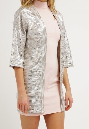Kobieta ubrana w jasnoróżową dopasowaną sukienkę z wysokim dekoltem oraz błyszczącą srebrną kurtkę z otwartym przodem i rękawami do połowy łokcia.