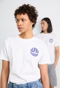 Baltie baltas kokvilnas T-krekliņš ar apaļu, zilu "ALPHA INDUSTRIES" logotipu kreisajā krūtī. Vienkāršs dizains ar īsām piedurknēm.