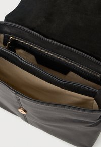 Μαύρη δερμάτινη τσάντα χειρός με σουέτ επάνω μέρος, διαθέτει τσεπάκι με φερμουάρ και καφέ φόδρα. Διακοσμημένη με μεταλλικό διακοσμητικό στο μπροστινό μέρος.