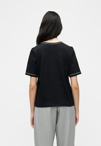 Γυναίκα με μακριά σκούρα μαλλιά που φοράει μαύρο τοπ με κοντά μανίκια και λευκή επένδυση, καθώς και ανοιχτό γκρι παντελόνι με λεπτές ρίγες, στέκεται γυρισμένη με την πλάτη.