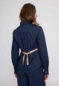 Tengerészkék gombos ing szabott hátrésszel és arany szatén szalaggal megkötve a deréknál, puha textúrájú és hosszú ujjú.