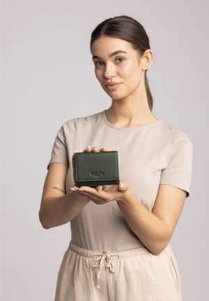 Green leather wallet with a textured finish, featuring a pressed logo. Compact rectangular shape held by a person.