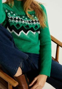 Femme assise sur une chaise en bois portant un pull à motifs verts, un jean bleu foncé et des bottines brunes.