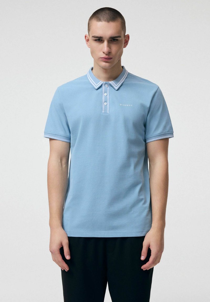 Młody mężczyzna stoi, ubrany w jasnoniebieską koszulkę polo z białą lamówką na kołnierzyku i rękawach, w połączeniu z czarnymi spodniami.