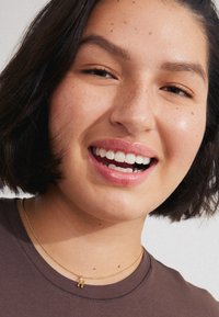 Prim-plan al unei persoane zâmbind, îmbrăcată cu un tricou maro și o brățară fină din aur cu un pandantiv în formă de literă "H". Pistruii sunt vizibili.