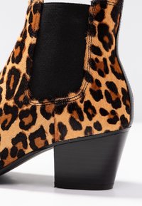 Leopardikuvioinen nilkkasaapas, jossa on tyylikäs musta kuminauhapaneeli ja teksturoitu vaaleanruskea ja musta turkispinta, korostettuna massiivisella mustalla korolla.