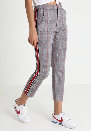 Femme portant un pantalon court à carreaux gris avec des bandes latérales rouges et noires, ainsi que des baskets blanches Nike avec un swoosh rouge.