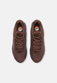 Brown athletic shoes with a textured mesh upper, featuring round laces and cushioned soles. Circular logo embedded on the tongue.