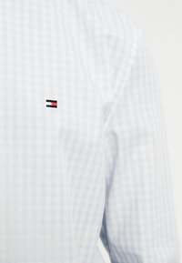 Μακρυμάνικο πουκάμισο γαλάζιο και λευκό καρό με μικρό ορθογώνιο κόκκινο, λευκό και μαύρο λογότυπο κεντημένο στο στήθος.