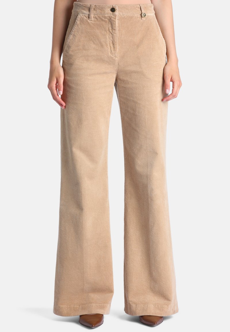 Pantalon large en velours côtelé beige porté par une personne debout, avec bouton et poches visibles, associé à des chaussures marron pointues.
