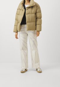 Beige vadderad jacka med hög krage, svart tröja under, vita byxor med subtilt mönster och grå sneakers med beige sulor.