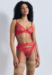 Σετ κόκκινων δαντελωτών εσωρούχων με διαφανές σουτιέν με κυματιστές άκρες, ταιριαστό ζαρτιέρα και κρεμ κάλτσες με λουλουδάτη δαντέλα.