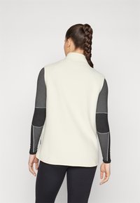 Cremfarbener Fleece-Westen mit hohem Kragen, der seitliche Einsätze in Dunkelgrau und kontrastierendem weißen Nähten aufweist. Schlichtes, elegantes Design.
