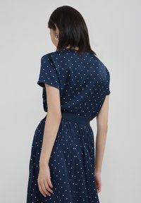 Abito blu navy con motivo a pois, maniche corte e vita aderente con gonna plissettata. Tessuto liscio con una leggera lucentezza.