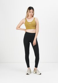 Sports bra in olive green, high waist black leggings, light sneakers. Smooth texture, adjustable straps, logo detail on the bra.