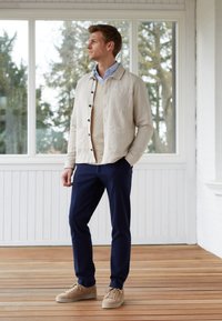 Giacca leggera beige chiaro sopra un maglione crema, abbinata a pantaloni blu navy e sneakers color sabbia. L'ambientazione presenta grandi finestre con vegetazione all'esterno.