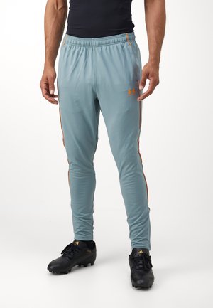 Mann trägt hellblaue Sporthose mit orangen Streifen und schwarzen Fußballschuhen, steht vor weißem Hintergrund.