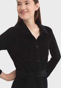 Donna che indossa un vestito nero con bottoni, dotato di taschini sul petto e una cintura legata in vita, sorridendo leggermente e guardando di lato.