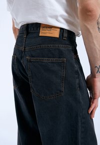 Black denim jeans featuring orange stitching, a leather tag with branding at the back, and two back pockets. The fit appears relaxed.