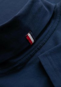 Tummansininen puuvillapaita, jossa on korkea kaulus. Paidassa on pieni kolmivärinen etiketti (punainen, valkoinen, sininen) pääntiellä ja ommeltuja saumoja.