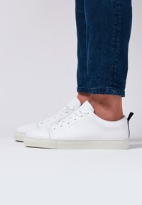 Sneakers bianchi in pelle con punta rotonda, suola piatta in gomma, sei fori per i lacci e design minimalista. Dettaglio della linguetta nera sul tallone. Indossati con jeans blu.