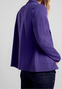 Fioletowa marynarka wykonana z miękkiego materiału; charakteryzuje się luźnym krojem, kołnierzem szalowym i bocznymi kieszeniami. Nosi się ją na ciemnych dżinsach.