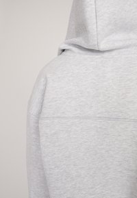 Grijze hoodie met een gestructureerde, zachte stof, voorzien van een voorzak en een capuchon met koord. De achterkant toont een naaddetail bij de schouders.