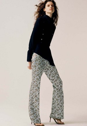 Femme aux longs cheveux ondulés porte un blazer asymétrique noir, un pantalon large à fleurs bleu clair et des sandales noires à talons hauts, debout de profil.