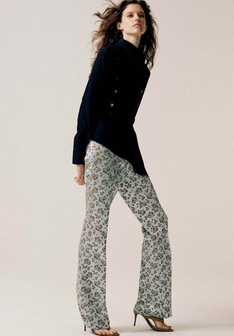 Femme aux longs cheveux ondulés porte un blazer asymétrique noir, un pantalon large à fleurs bleu clair et des sandales noires à talons hauts, debout de profil.