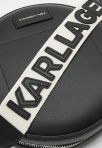 Borsa rotonda nera in pelle con una tracolla bianca che presenta lettere nere in rilievo che spelling "KARL LAGERFELD" e un dettaglio con zip.