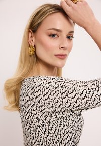 Czarna i biała sukienka w wzory z dzwonkowymi rękawami; modelka nosząca złote kolczyki z kwiatowymi zdobieniami; długie, proste włosy opadające na ramię.