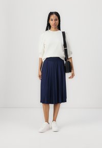 Light cream pullover with puff sleeves paired with a navy pleated skirt. White sneakers and a small black bag complete the look.
