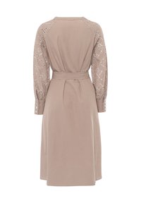 Beige jurk met een aansluitende taille en een klokvormige rok. Voorzien van geborduurde transparante mouwen met ronde uitsparingen en knoopmanchetten.