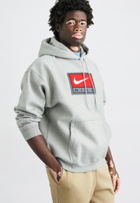 Γκρι φούτερ με κουκούλα, μπροστινή τσέπη και κορδόνια. Διαθέτει κόκκινο λογότυπο Nike σε ορθογώνιο πατς. Μαλακό υλικό και άνετη εφαρμογή.